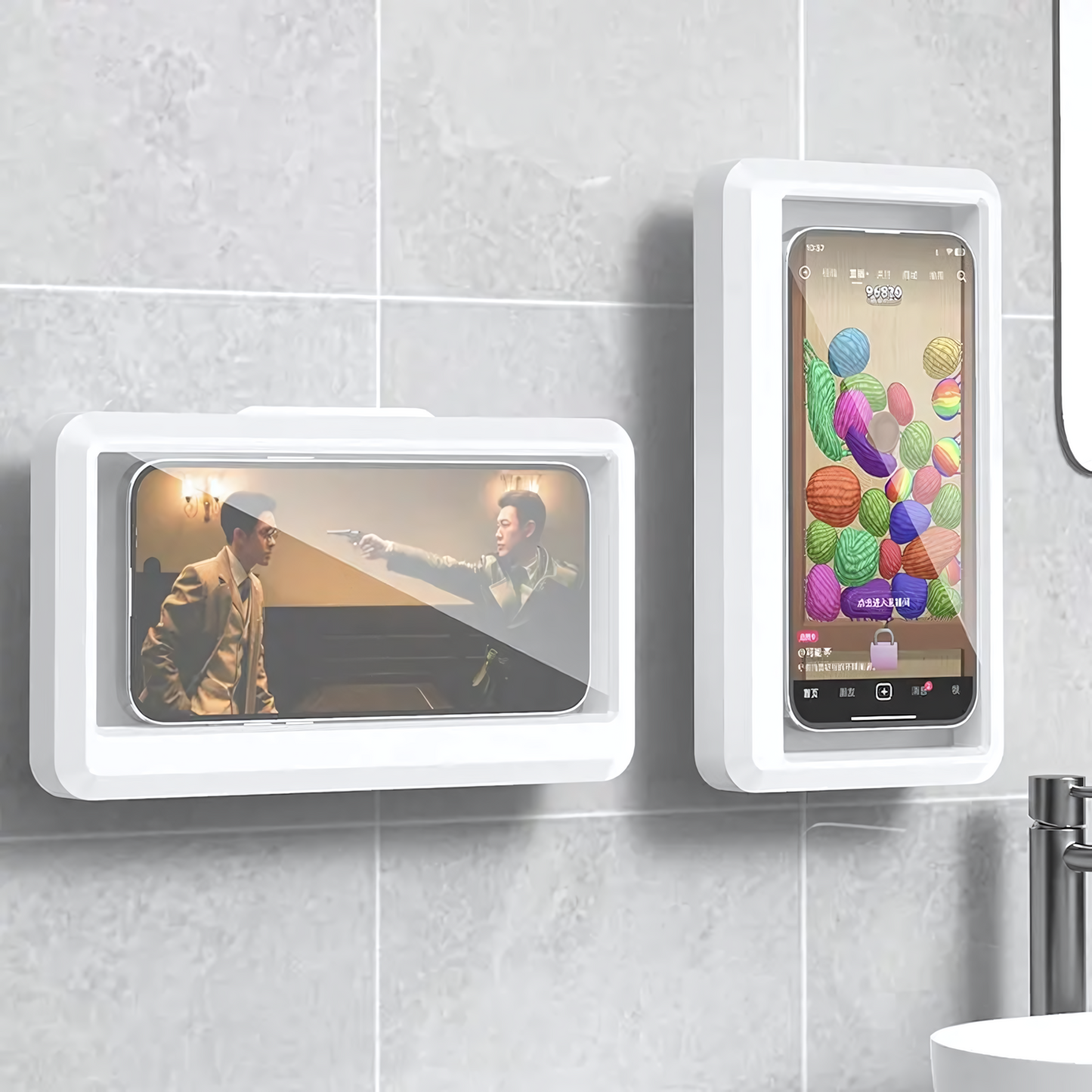 Un mur de salle de bain est orné de deux supports de téléphone étanches PhonoNest™ de Homesafetouch, dotés d'une étanchéité à 360° et de coques de protection en plastique. Le support de gauche présente un téléphone diffusant une scène de film avec deux hommes, tandis que celui de droite présente un jeu avec des pelotes de laine colorées. Un robinet argenté est partiellement visible en bas à droite.